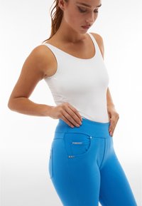 Pantaloni stretch blu con vita alta, dotati di una tasca laterale e accenti metallici decorativi. Il modello indossa un top bianco aderente.