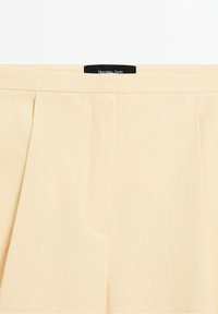 Pantalon en lin beige clair avec plis et une ceinture, montrant l'étiquette de la marque "Massimo Dutti" à l'intérieur de la ceinture.