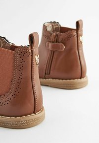 Bottines en cuir marron avec fermeture éclair latérale, panneaux latéraux élastiques, bords festonnés et détails perforés sur les côtés extérieurs.
