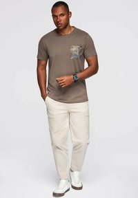 Šedé tričko s květinovou kapsou, spárované s béžovými cargo kalhotami a bílými teniskami. Osoba stojí vzpřímeně a rukou naznačuje vpřed.