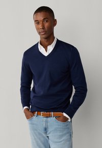 Mężczyzna w granatowym swetrze z dekoltem w serek na białej koszuli, jasnoniebieskich dżinsach i brązowym skórzanym pasku, stoi z rękami w kieszeniach.
