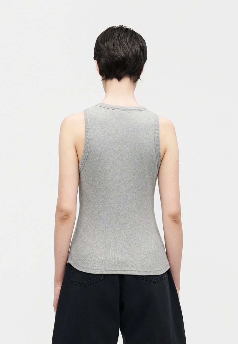 Personne aux cheveux courts et foncés, portant un haut gris en maille sans manches et un pantalon noir, debout de dos sur un fond blanc uni.