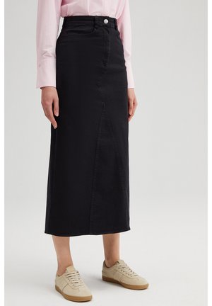 Vrouw draagt een zwarte midi-rok met hoge taille en vooraan een split, een lichtroze blouse ingestopt en beige veter sneakers, staand op een witte vloer.