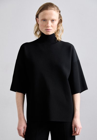 Haut noir surdimensionné à col roulé avec des manches courtes, texture lisse et silhouette structurée, porté avec un pantalon noir assorti.