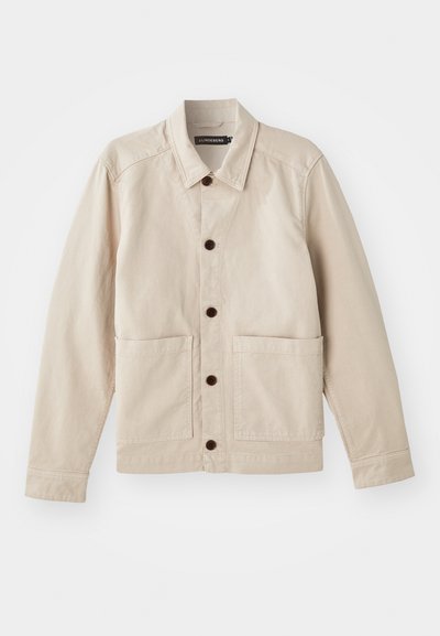 Chaqueta de algodón beige con cierre de botones en la parte delantera, cuello puntiagudo, dos bolsillos frontales y botones marrones; presenta un diseño estructurado y casual.