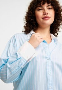 Ljusblå randig skjorta med knäppning fram, vita manschetter och en avslappnad passform med en slät textur.