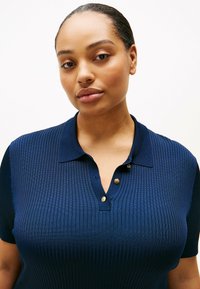Marineblaues, geripptes Poloshirt mit klassischem Kragen, kurzen Ärmeln und goldenen Knopf-Akzenten am Ausschnitt für einen strukturierten Look.
