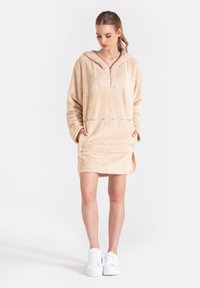 Beżowa sukienka z polaru z kapturem, z kieszenią z przodu, na pół zamku i długimi rękawami; miękka tekstura i luźny krój. Noszona z białymi trampkami.