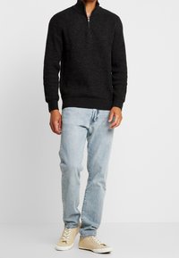Schwarzer Strick-Zipfelpullover mit gerippter Textur, kombiniert mit hellblauen Jeans und beigen Sneakers. Die strukturierte Gestaltung betont den Komfort.