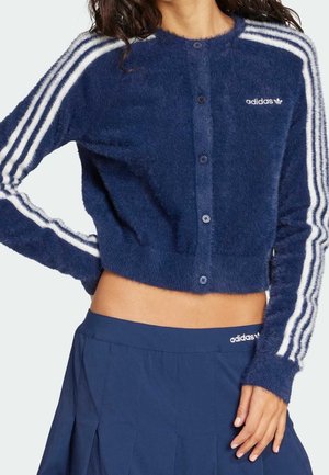 Granatowy, miękki kardigan na guziki z białymi paskami Adidas na rękawach, zestawiony z granatową spódnicą Adidas, noszony przez osobę.
