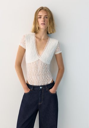 Kvinde med skulderlangt blondt hår iført en gennemsigtig hvid blondebluse med korte ærmer og mørke jeans med høj talje, stående med hænderne i lommerne.