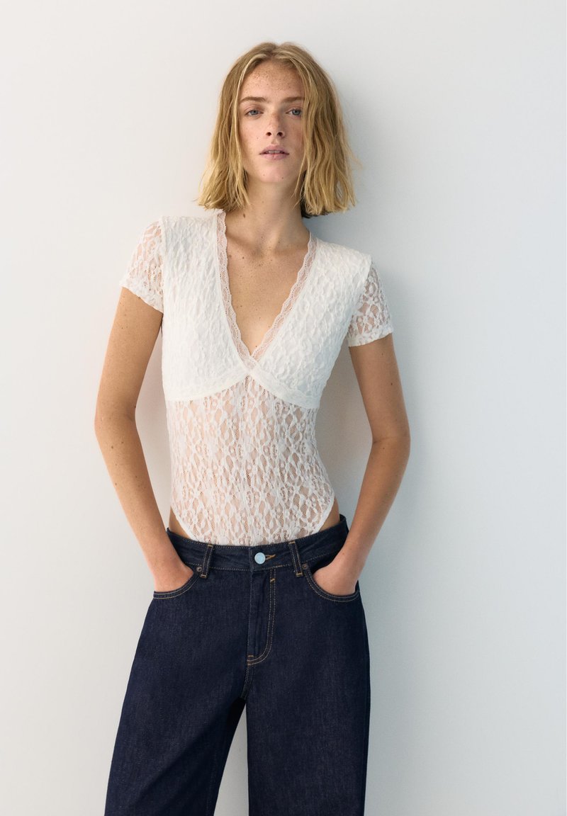 Femme aux cheveux blonds mi-longs portant un haut blanc en dentelle transparente à manches courtes et un jean taille haute foncé, debout les mains dans les poches.