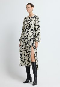 Largo vestido fluido en crema con estampado floral negro, con mangas largas, escote fruncido y abertura lateral. Combinado con botas negras hasta la rodilla.