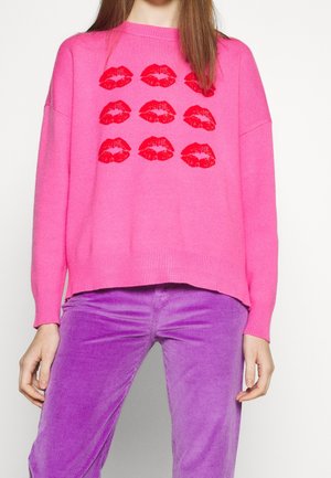 Personne portant un pull rose vif avec des imprimés de lèvres rouges et un pantalon en velours côtelé violet, debout devant un fond uni.