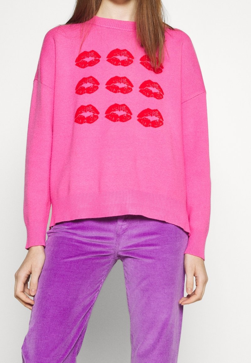 Personne portant un pull rose vif avec des imprimés de lèvres rouges et un pantalon en velours côtelé violet, debout devant un fond uni.