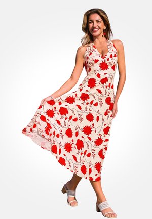 Kobieta uśmiechająca się, ubrana w białą sukienkę bez rękawów z czerwonym kwiatowym wzorem, trzymająca jedną stronę sukienki i nosząca białe sandały na słupku.