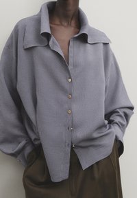 Chemise à rayures bleues et blanches avec un grand col, texture douce et boutons en or, portée sur un pantalon sombre.