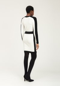 Abito aderente bianco e nero con maniche lunghe, texture a coste, dettagli laterali e colletto alto, abbinato a collant neri fino alla coscia e tacchi.