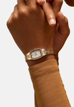 Silver and gold-tone wristwatch with rectangular face, white dial, and crystal accents. Worn on a hand with brown fabric sleeve.