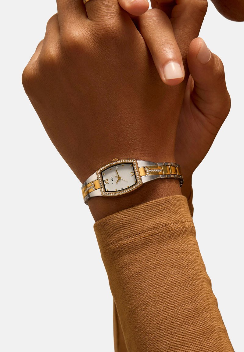 Montre-bracelet en argent et or avec un cadran rectangulaire, un cadran blanc et des accents en cristal. Portée sur une main avec une manche en tissu brun.
