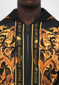 Haut à zip noir avec un motif floral baroque doré et un texte vertical indiquant "Versace Jeans Couture" sur les panneaux avant.