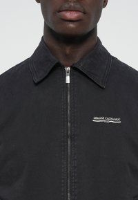 Black zip-up jacket, cotton blend, with a pointed collar. Features silver zipper and "Armani Exchange 1991" logo embroidered on chest.