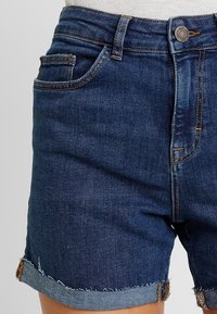 Tmavě modré džínové kraťasy s roztřepeným lemem, klasickým pěti-kapsovým designem a kontrastním prošitím na pase a kapsách.