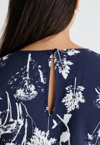 Marinefarbener Stoffoberteil mit weißem botanischen Druck, Rückenschlitz mit Knopfdetail und glatter Textur.