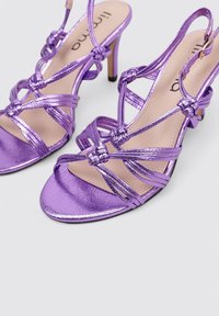 Sandalias de tacón moradas con acabado metálico, tiras entrelazadas con detalles de nudos y un tacón estrecho. Textura interior suave.