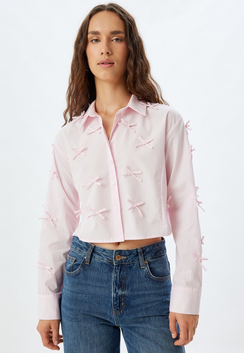 Camisa corta rosa de tejido ligero, con botonera frontal, cuello puntiagudo y lazos decorativos en las mangas. Combinada con vaqueros azules.