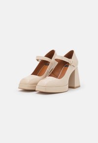 Beige patenterade läder Mary Jane plattformsskor med breda blockklackar och justerbara spännen på en vit bakgrund.