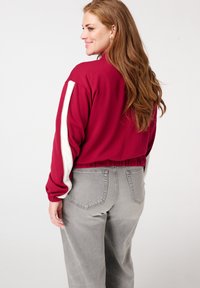 Sweatshirt bordeaux court avec des rayures blanches sur les côtés, ourlet élastique et coupe loose. Porté avec un jean taille haute gris clair.