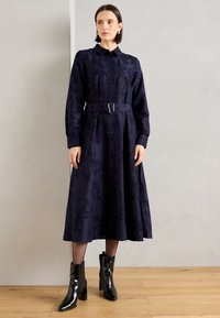 Marineblauw knielang shirtjurkje met lange mouwen, een ceintuur in de taille, bloemenmotief en een volle rok. Gemaakt met zwart enkellaarsjes met een hak.