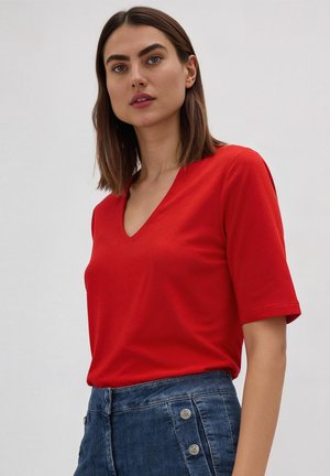 Frau mit schulterlangem braunem Haar, die ein rotes V-Ausschnitt-Kurzarmshirt und hoch taillierte blaue Jeans mit Knopfdetails trägt.