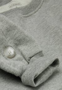 Lys grå sweater med ribbet hals og manchetter, med en stor perleknapdetalje på den optrukne ærme. Blød stof tekstur.