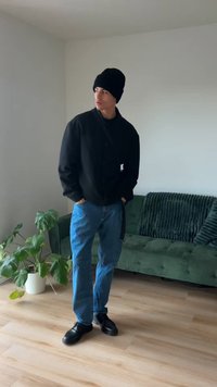 Mustast nuppudega jakk, sinised teksapüksid, mustad kingad ja talvemüts; seisab toas, kus on puidust põrand ja roheline pehme diivan.