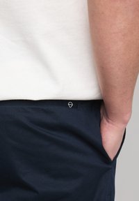 Shorts navy avec une texture lisse, présentant un petit logo triangulaire sur la ceinture, et une main dans la poche avant.