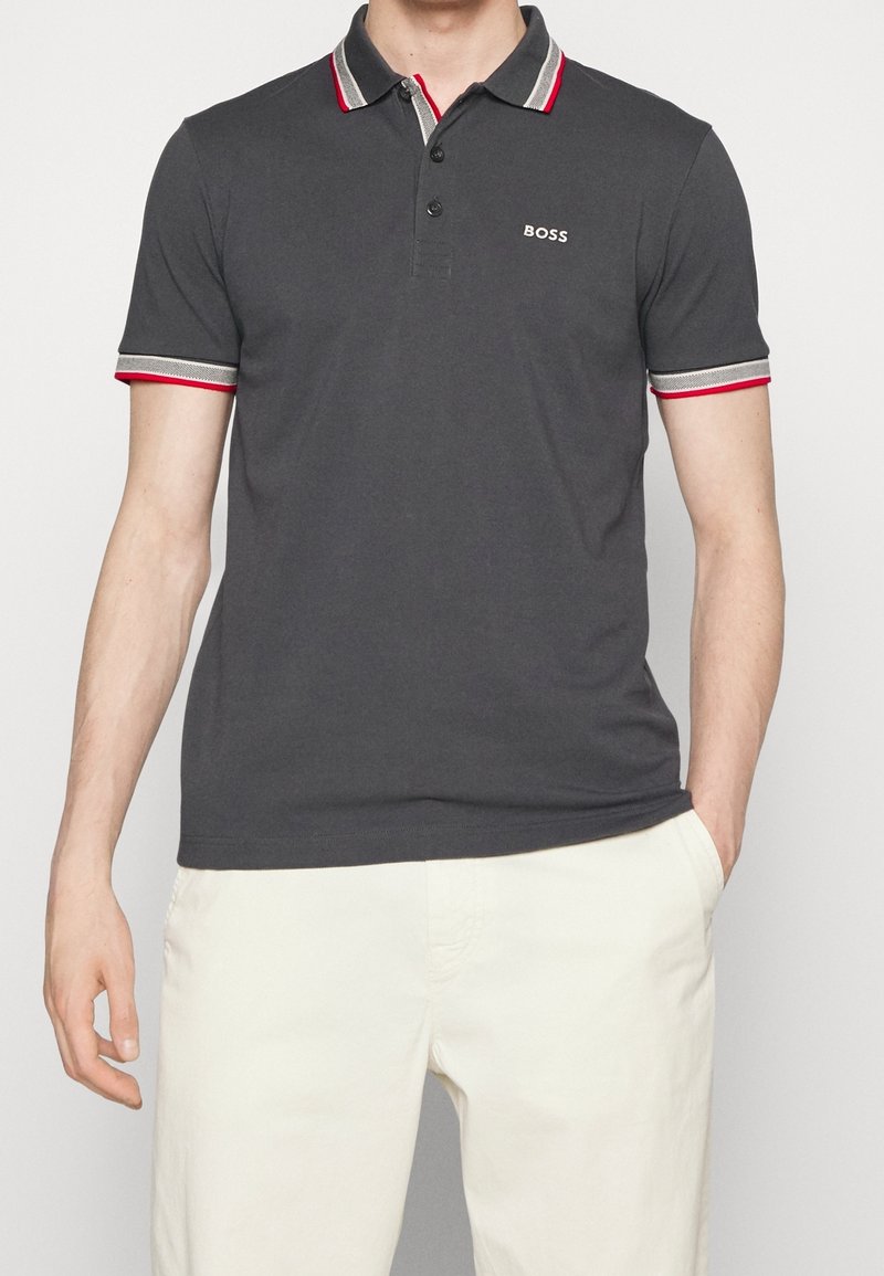 Polo gris foncé avec un col classique, présentant des accents rayés rouges et gris sur les manches et un petit logo brodé sur la poitrine.