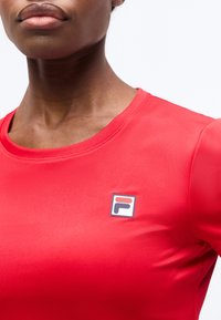 Κοντινό πλάνο ενός ατόμου που φοράει μια φωτεινή κόκκινη μπλούζα με ένα μικρό λογότυπο FILA στην αριστερή πλευρά του στήθους.