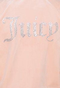 Rosa klädesplagg i sammet med ordet "Juicy" i stora silvriga strassstenar, visat över ryggen i ett kursivt typsnitt.