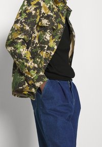 Giacca mimetica con motivi verdi, gialli e marroni, con chiusura a bottoni, abbinata a una camicia nera e pantaloni in denim blu.