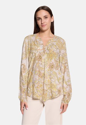 Vrouw met lang bruin haar die een lichtbeige blouse draagt met gele en gouden bloemige paisley-patronen en crèmekleurige broek.