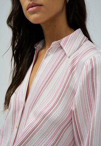 Blusa a rayas con rayas verticales en rosa y blanco con acentos metálicos brillantes. Diseño con cuello y cierre de botón en el escote.