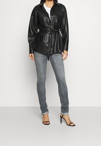 Černá kožená košile s páskem, zapínáním na knoflíky a dvěma náprsními kapsami, doplněná světle šedými úzkými džínami a černými sandály na podpatku.