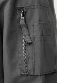 Κλείσιμο τσέπης από γκρίζο ύφασμα με ορατές ραφές και μεταλλικό φερμουάρ που φέρει λογότυπο δέντρου στην ταμπέλα έλξης.