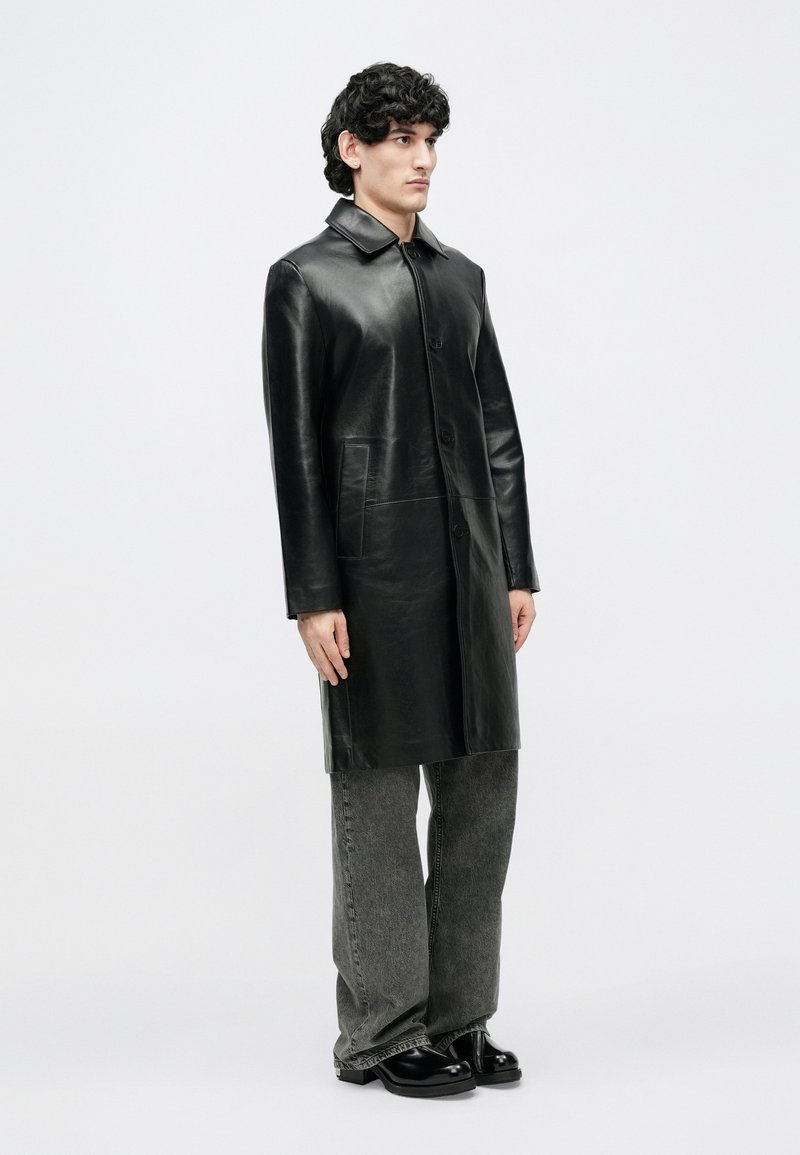 Homme debout portant un long manteau en cuir noir, un jean gris ample et des chaussures noires, sur un fond blanc uni.