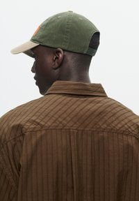 Bruine gestreepte shirt met een omklapbare kraag, gecombineerd met een olijfgroene pet met een beige rand en een verstelbare strap.