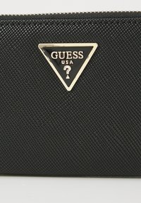 Mustra mustrahall, millel on kolmnurkne kuldne ja must logo plaat, millel on kirjas "GUESS USA" ja küsimärgi disain. Lukuga sulgemine.