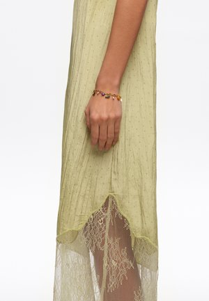 Mano izquierda descansando a un lado, con una pulsera de dijes dorada, vestida con un vestido verde claro con dobladillo de encaje transparente y textura de puntos.