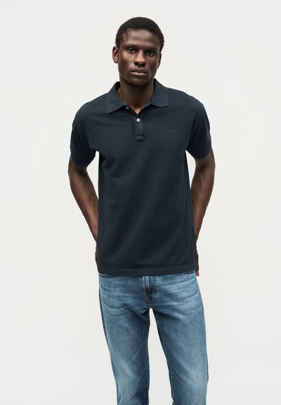 Νέος άνδρας που στέκεται με τα χέρια πίσω από την πλάτη, φοράει σκούρο μπλε πόλο και ανοιχτόχρωμο τζιν μπροστά σε ένα απλό φωτεινό φόντο.